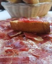 Plato de jamón ibérico con pan y bebidas en la mesa, cesta con pan al fondo.