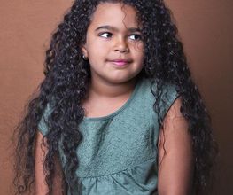 Niña de cabello rizado, con camiseta verde y pantalón amarillo, mirando hacia arriba.
