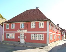 Rotes Gasthaus mit Satteldach an einer Straßenecke, weiß gerahmte Fenster, klare Wetterlage.