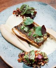 Prato gourmet com peixe grelhado, purê e guarnições coloridas em prato cinza sobre mesa de madeira.