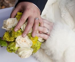 Mani di sposi con fedi nuziali sopra un bouquet di rose bianche e verdi.