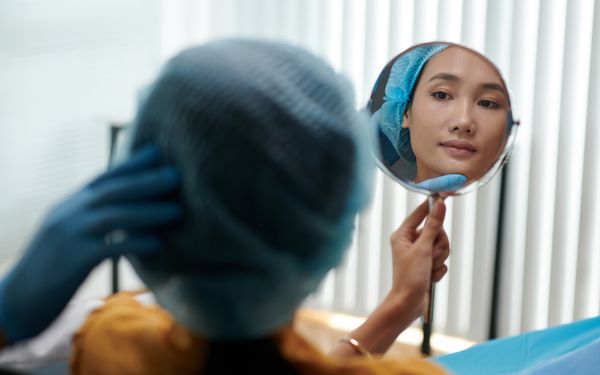 Eine Frau mit Haarnetzt, hält sich einen Handspiegel vor ihr Gesicht, während der Schönheitschirurg erklärt was er verändern würde.