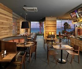 Restaurante moderno de madera con vista al mar, mesas vacías y pizarras decorativas.