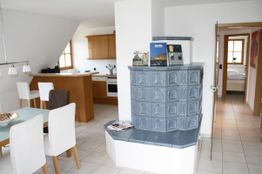 Helle Küche und Essbereich mit blauem Kaminofen, Esstisch und Blick in ein Schlafzimmer.