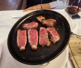 Carne asada sobre plancha negra, junto a copa de vino tinto y tabla de cortar en mesa blanca.