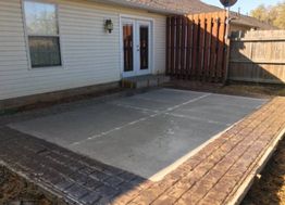 Concrete patio with a brick border in a backyard beside a house with white siding.