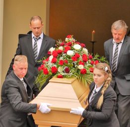 Menschen in Anzügen tragen einen geschmückten Sarg bei einer Trauerfeier.