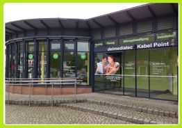 Gebäude mit der Beschriftung "delmediatec Kabel Point", Glastüren und Plakaten an der Fassade.