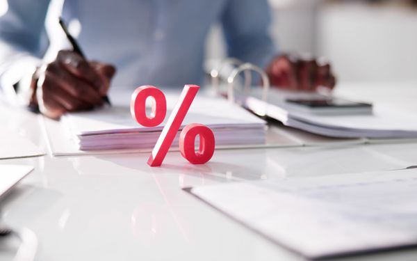 Un hombre escribe algo en un documento que está archivado en una carpeta frente a él. Delante de la carpeta hay un símbolo de porcentaje rojo que representa el IVA.