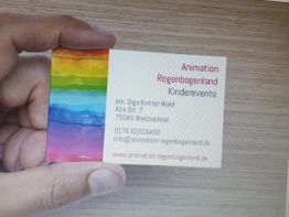 Hand hält Visitenkarte mit Regenbogenmotiv und Kontaktdaten für Kinderevents.