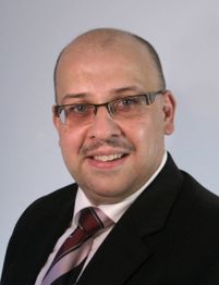 Ein lächelnder Mann mit Brille und Anzug posiert vor einem neutralen Hintergrund.