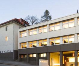 Moderne Gebäudeansicht bei Dämmerung, mit beleuchteten Fenstern und Bäumen im Hintergrund.