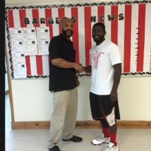 Two men shake hands in front of a bulletin board with "Barber's News" written on it.