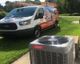 HVAC van parked beside an outdoor air conditioning unit on green grass.