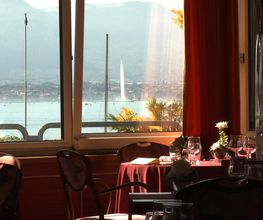 Blick aus einem Restaurantfenster auf einen See mit Bergkulisse im Hintergrund, Tische im Vordergrund.