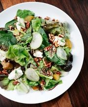 Prato de salada verde com queijo, nozes e pepino sobre mesa de madeira escura.