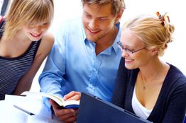 Drei Personen lernen zusammen mit einem Buch und Laptop. Alle lächeln und wirken konzentriert.