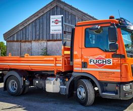 Ein orangefarbener LKW mit der Aufschrift "Fuchs" steht vor einer Holzscheune.