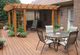 Wooden deck with pergola, patio table, chairs, potted plants, and brick wall background.