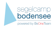 Logo mit blauem Dreieck und Text "segelcamp bodensee powered by BeOneTeam" in verschiedenen Farben.