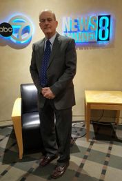 Man in a suit standing by ABC 7 News Channel 8 sign, next to a chair and table.