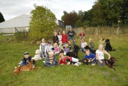 Kinder und Hunde sitzen zusammen auf einer Wiese vor Büschen und einem Zaun.