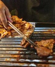 Pessoa grelhando carnes e linguiças em uma churrasqueira, usando pegador de metal.