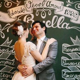 Noivos abraçados em frente a parede decorada com palavras como "amor" e "amizade".