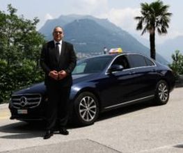 Ein Mann steht im Anzug neben einem schwarzen Mercedes-Benz vor Bergkulisse und Palmen.
