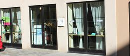 Fensterfront eines Gebäudes mit Vorhängen und Schildern, daneben Pflastersteine und ein rotes Auto.