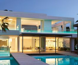 Moderne Villa mit Glasfronten, beleuchtet bei Dämmerung, mit Pool und Palmen im Garten.
