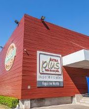 Fachada de supermercado com paredes vermelhas e logotipo "Plus" em destaque sob céu azul.