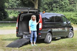 Frau mit blauem Shirt steht an einem schwarzen Fahrdienst-Van mit geöffneter Heckklappe und Rampe.