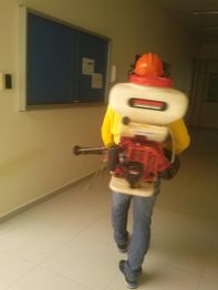 Pessoa com capacete e pulverizador nas costas, caminhando em corredor de prédio.