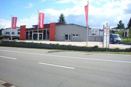 Modernes Gebäude mit roten Akzenten, Werbeflaggen vor der Fassade, sonniger Tag.