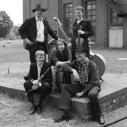 Gruppe von fünf Musikern mit Instrumenten posiert draußen neben einem Backsteingebäude.