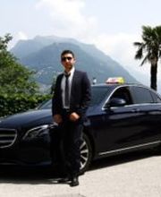 Ein Mann in Anzug steht vor einem schwarzen Mercedes auf einer Straße mit Berg- und Palmenhintergrund.