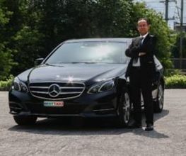 Mann in Anzug steht mit verschränkten Armen vor einem schwarzen Mercedes auf Parkplatz.