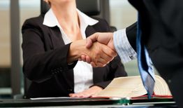 Dos personas dándose la mano en una oficina, con un libro abierto en la mesa.