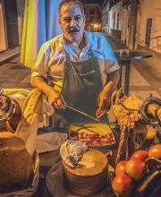 Hombre en delantal con mesas de embutidos, queso y verduras en una calle iluminada.