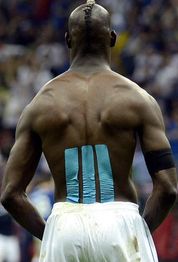 Uomo di spalle indossa pantaloni sportivi bianchi, cerotti blu sulla schiena, folla sullo sfondo.