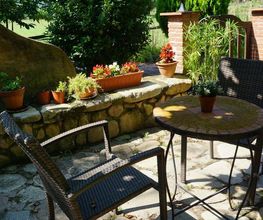 Patio con sillas, mesa y macetas, rodeado de vegetación con montañas al fondo en un día soleado.