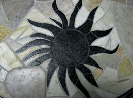 Schwarze Sonnenform auf Steinmosaikboden aus beigen und grauen Fliesen.