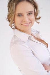 Eine lächelnde Frau in weißem Hemd vor hellem Hintergrund, mit verschränkten Armen.