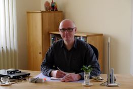 Ein Mann sitzt an einem Schreibtisch mit Pflanzen und Büchern im Hintergrund.