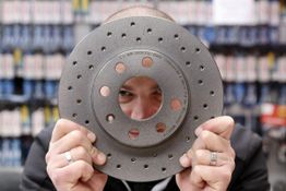 Person hält Bremsscheibe mit Gesicht dahinter, schaut durch die Löcher. Regal im Hintergrund.