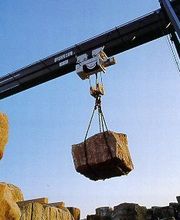 Kran hebt großen Steinblock gegen blauen Himmel. Weitere Steine liegen links unten.