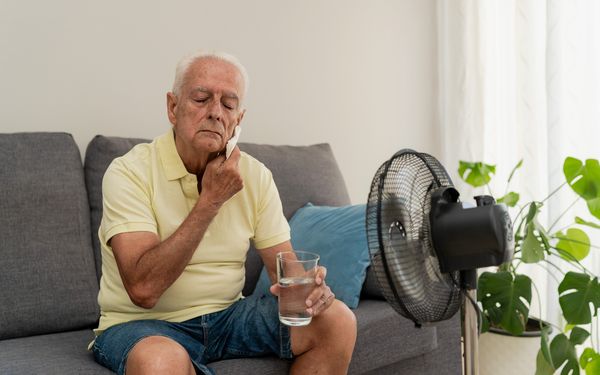 Älterer Mann sitzt mit einem Glas Wasser vor einem Ventilator und wischt sich bei Hitze das Gesicht ab.