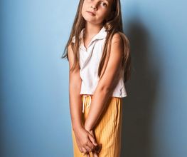 Niña de pie en fondo azul, con camisa blanca y pantalones amarillos, mirando a la cámara.