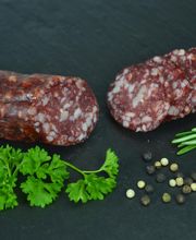 Salami, Pfefferkörner, Petersilie und Rosmarin auf Schieferplatte vor schwarzem Hintergrund.